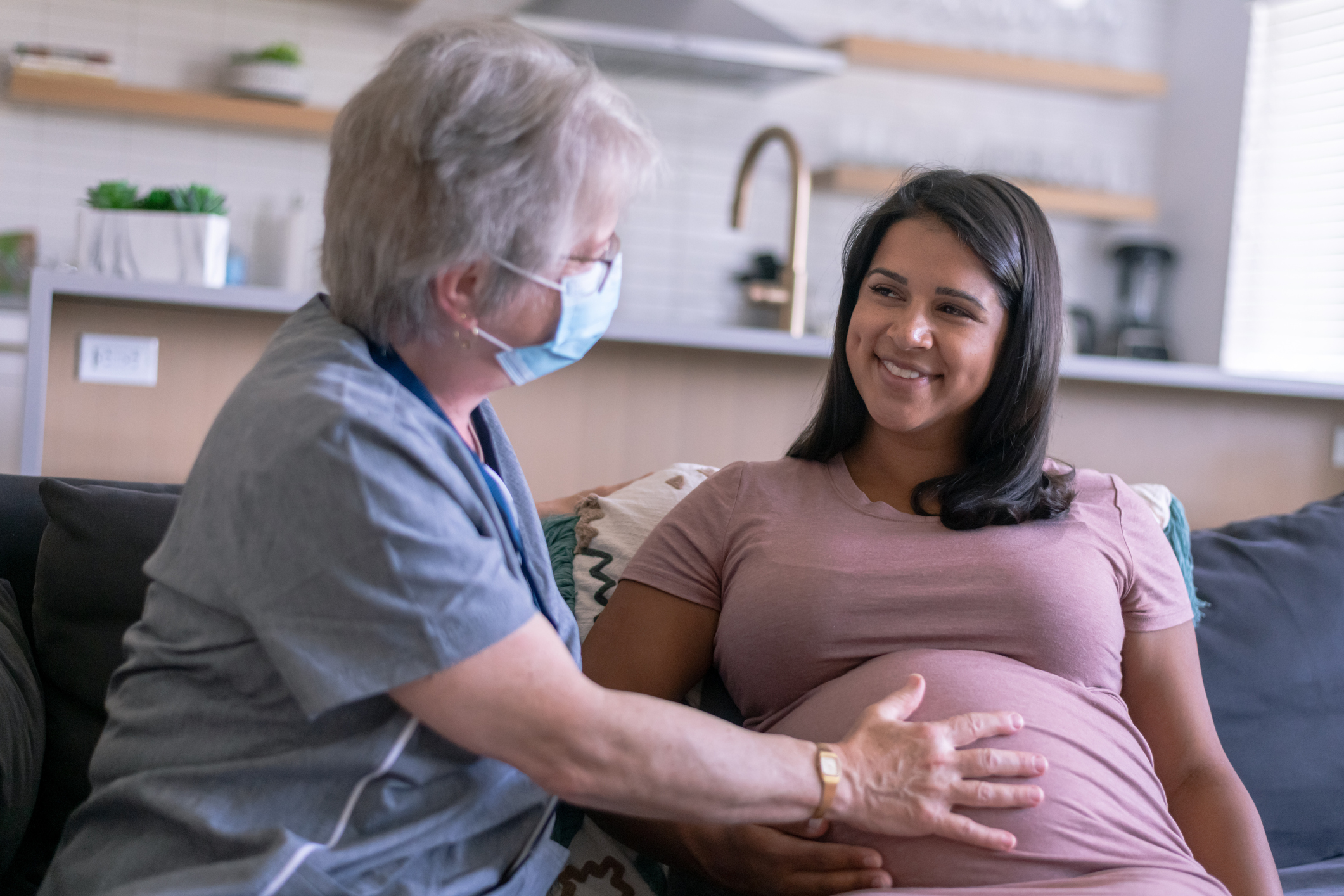 A beautiful young adult ethnic woman is pregnant and having an in-home medical check up with her nurse. The nurse is an older caucasian woman. She is checking the woman's belly and has her hand placed on her stomach while wearing a surgical mask because of COVID-19.