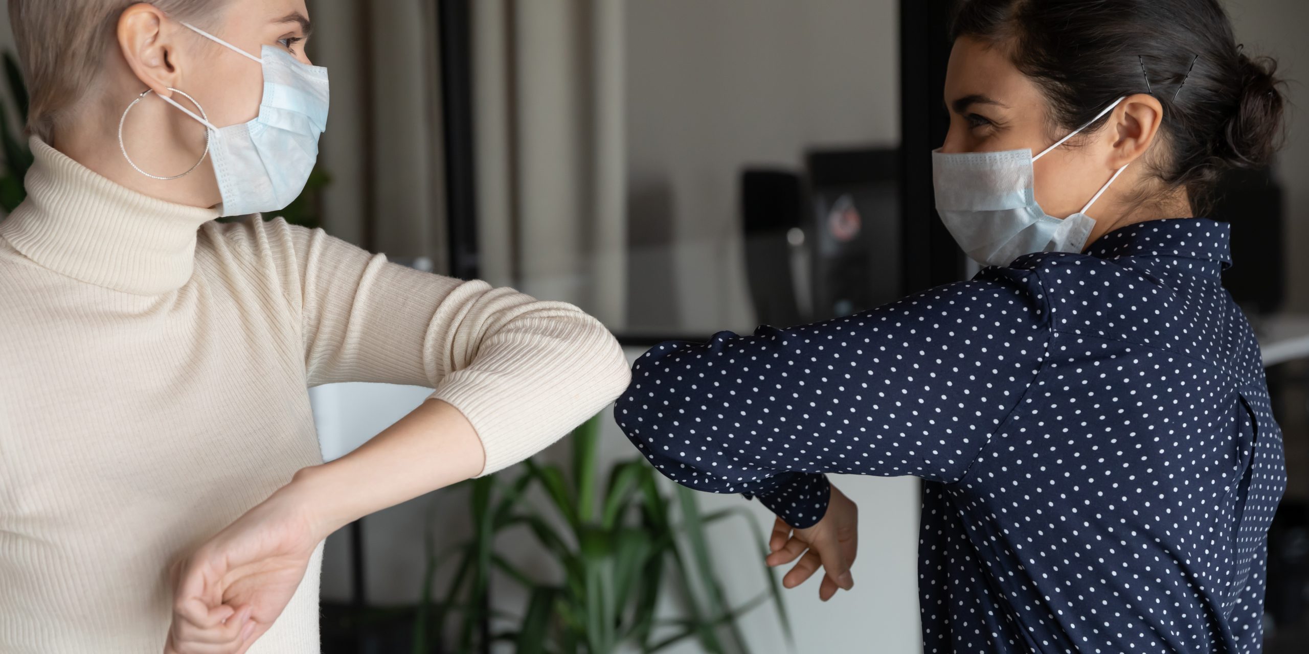 Smiling young healthy mixed race female colleagues wearing facial medical masks greeting each other by bumping elbows gesture at workplace keeping social distance, preventing spreading covid19 virus.