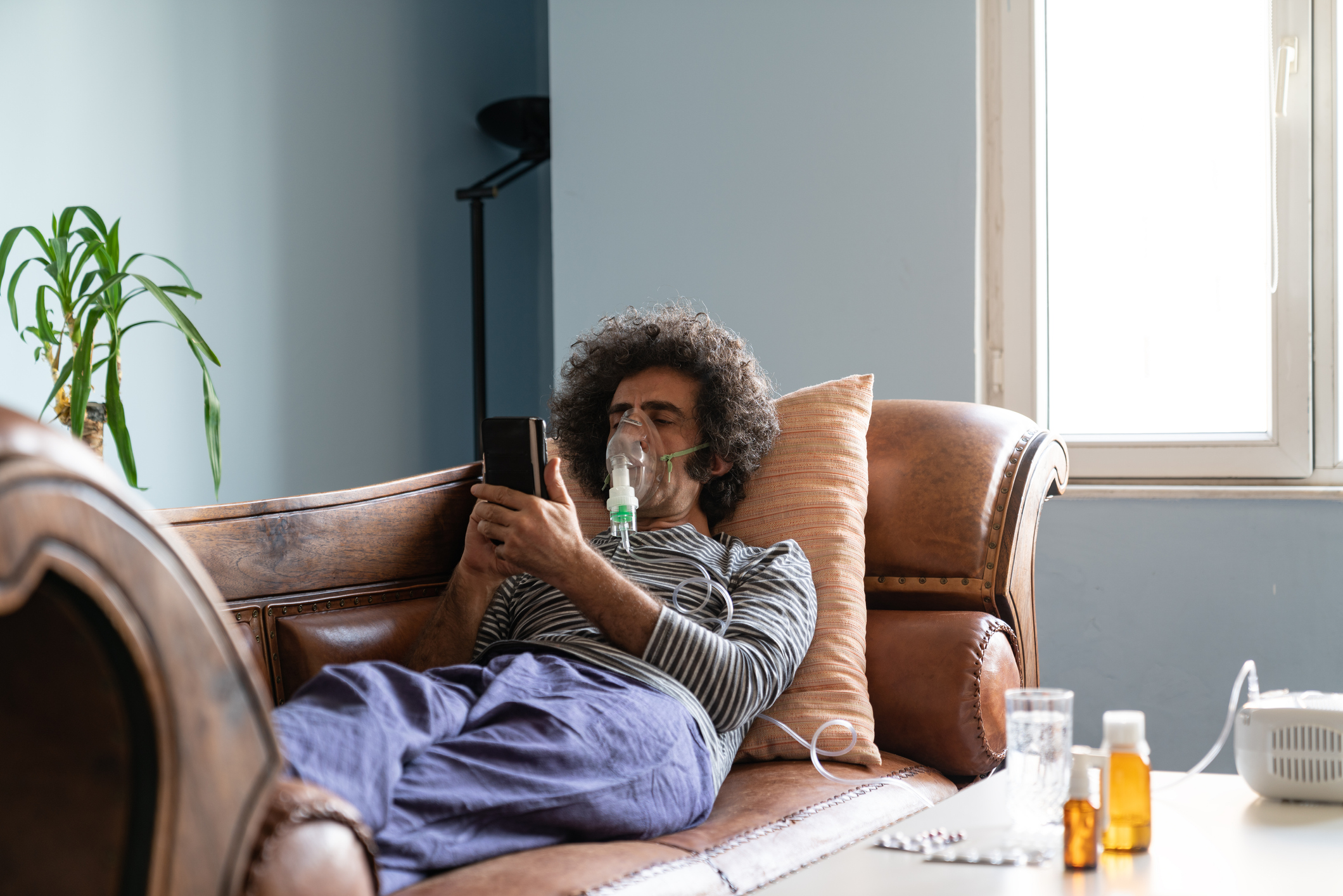Mature adult man lying down on sofa in living room and using nebulizer during coronavirus pandemic. He is wearing sleepwear and using smartphone for communication. Shot indoor with a full frame mirrorless camera.