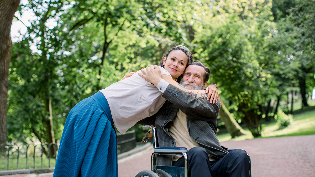 Disabled people, palliative care. Lifestyle summer portrait of senior bearded man in wheelchair hugging his young pretty hipster granddaughter, walking in park outdoors.