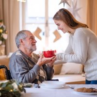 Young woman giving cup of tea to senior grandfather in wheelchair indoors at home at Christmas.