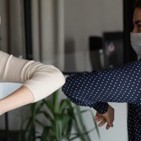 Smiling young healthy mixed race female colleagues wearing facial medical masks greeting each other by bumping elbows gesture at workplace keeping social distance, preventing spreading covid19 virus.