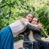 Disabled people, palliative care. Lifestyle summer portrait of senior bearded man in wheelchair hugging his young pretty hipster granddaughter, walking in park outdoors.