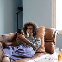 Mature adult man lying down on sofa in living room and using nebulizer during coronavirus pandemic. He is wearing sleepwear and using smartphone for communication. Shot indoor with a full frame mirrorless camera.