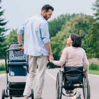 happy man holding hand of disabled wife while walking with baby carriage in park together