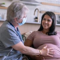 A beautiful young adult ethnic woman is pregnant and having an in-home medical check up with her nurse. The nurse is an older caucasian woman. She is checking the woman's belly and has her hand placed on her stomach while wearing a surgical mask because of COVID-19.