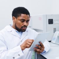 Serious concentrated young black medical scientist with beard sitting at desk and using digital tablet while analyzing data in laboratory